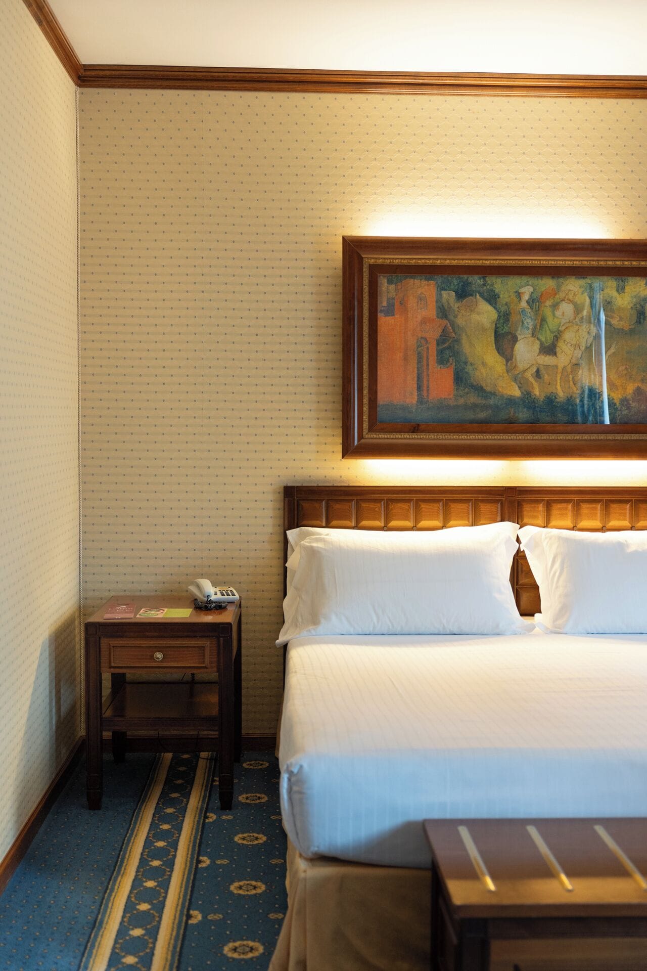Una habitación de hotel con una cama, una mesita de noche con un teléfono, un cuadro sobre la cama y una alfombra estampada.