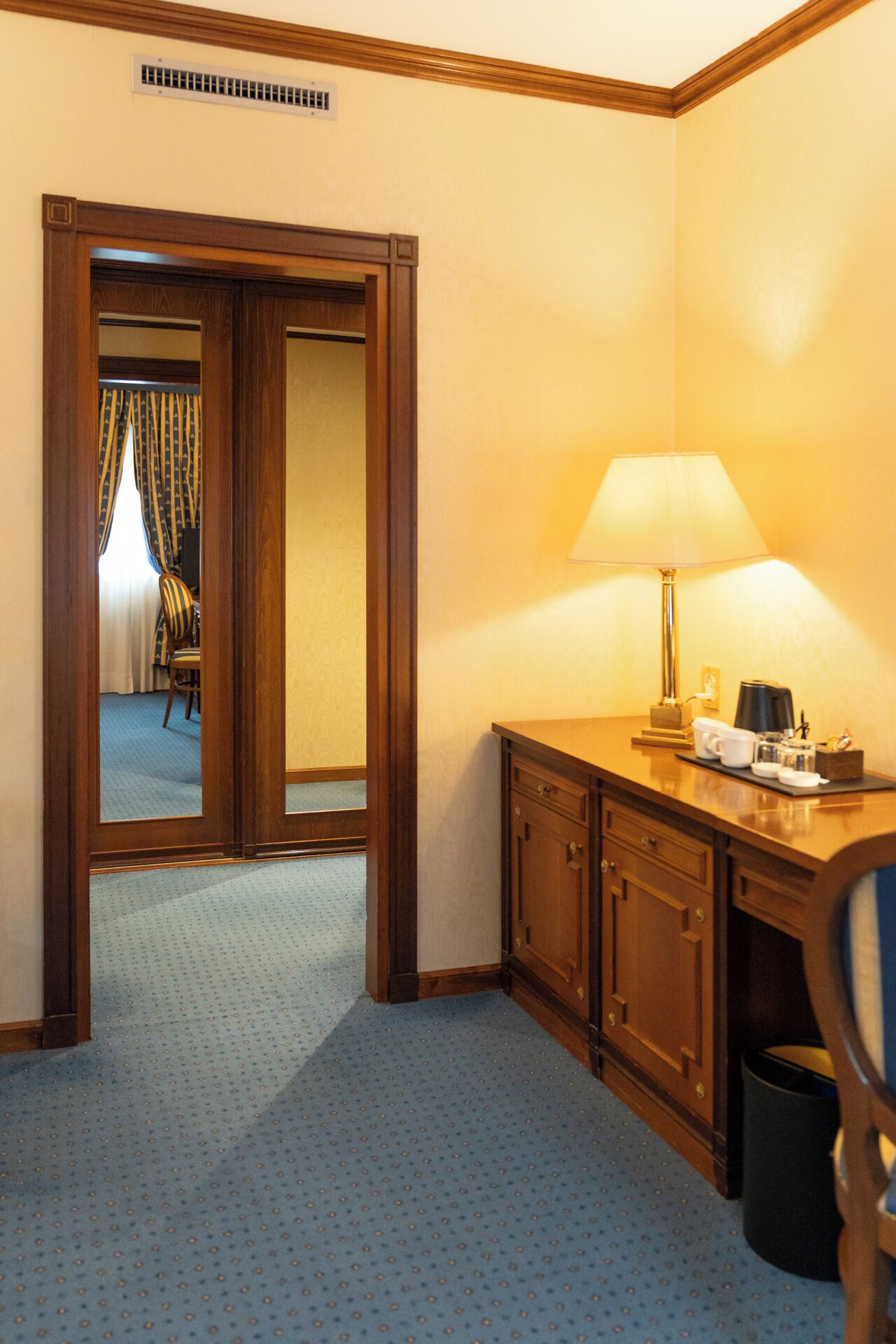 A hotel room with a wooden desk, a lamp, a chair, a mirror, and a door leading to another room.
