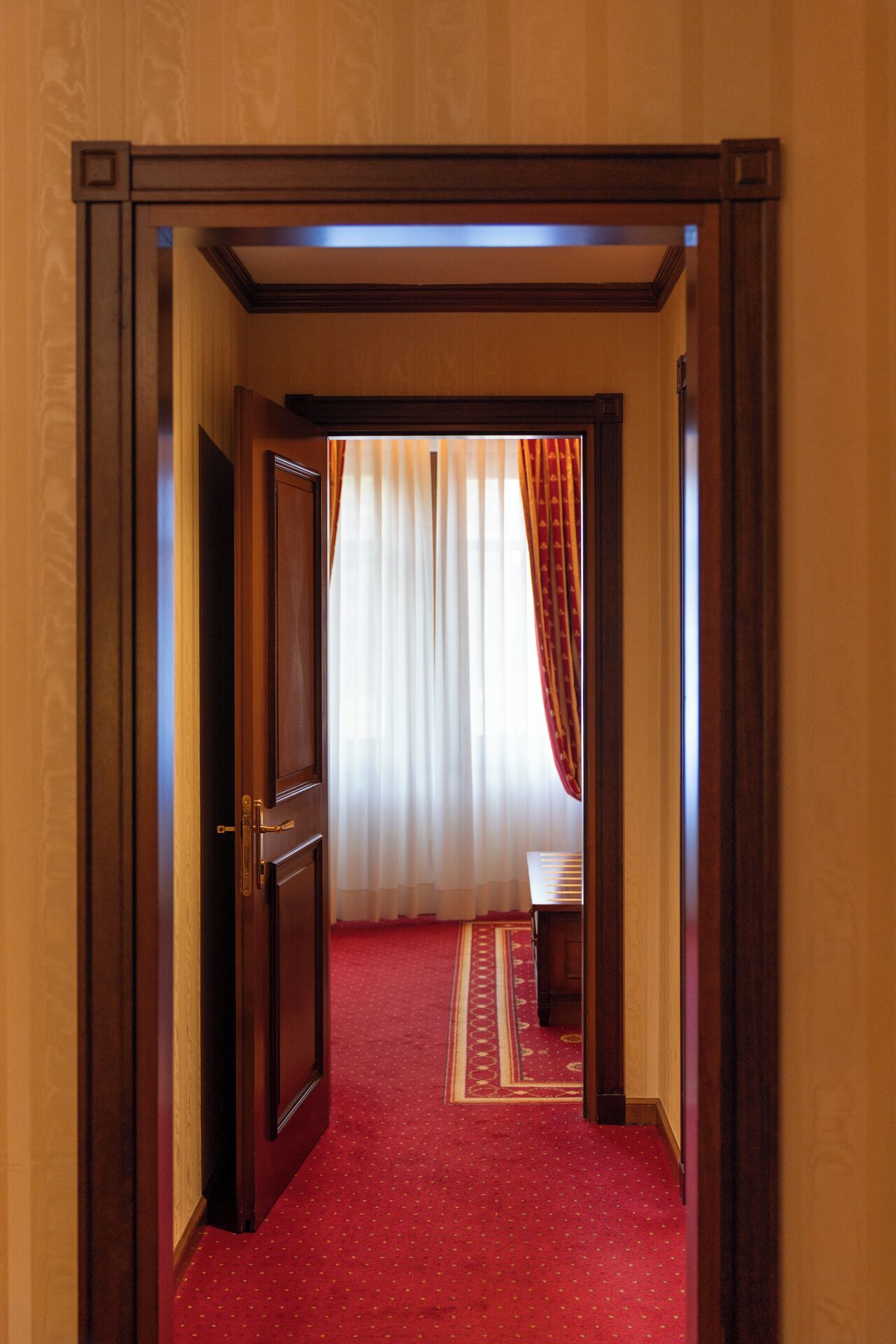 A hallway with a red carpet leading to a room with a bed and a window with curtains.