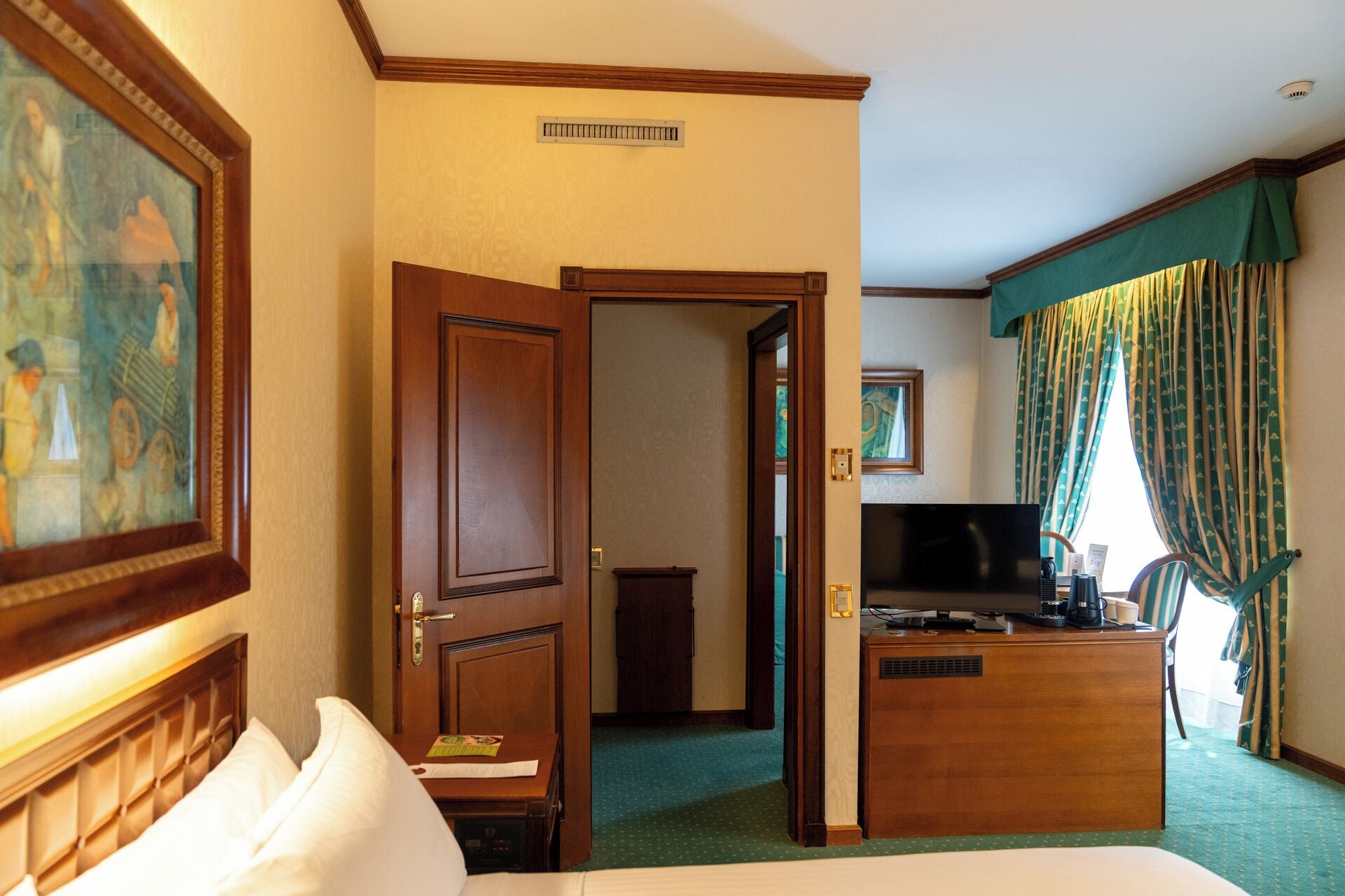 Habitación de hotel con cama, escritorio con televisor, puerta y un cuadro en la pared.