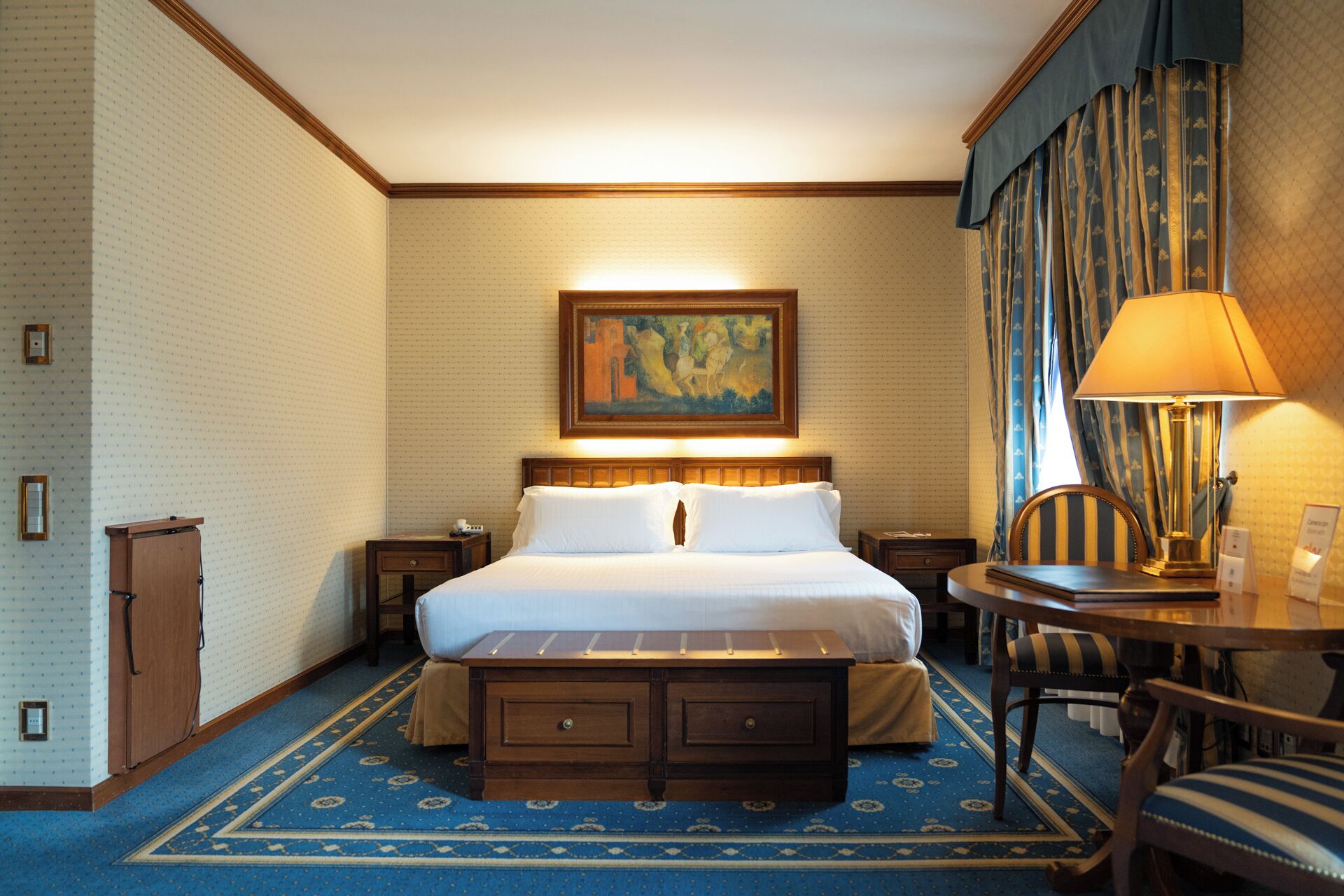 A hotel room with a bed, a desk, a chair, a lamp, a painting on the wall, and a patterned carpet.