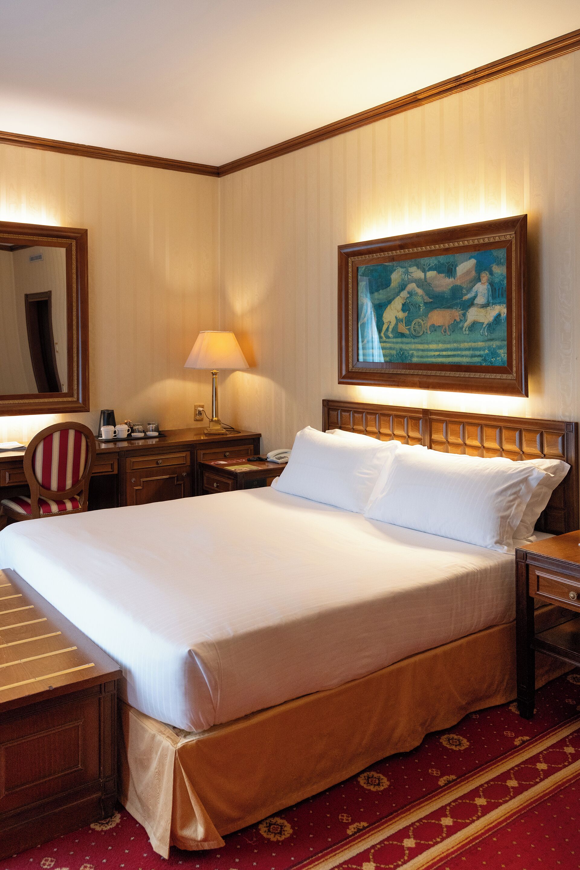 A hotel room with a large bed, a desk, a chair, a nightstand, a lamp, a painting, and a carpet.