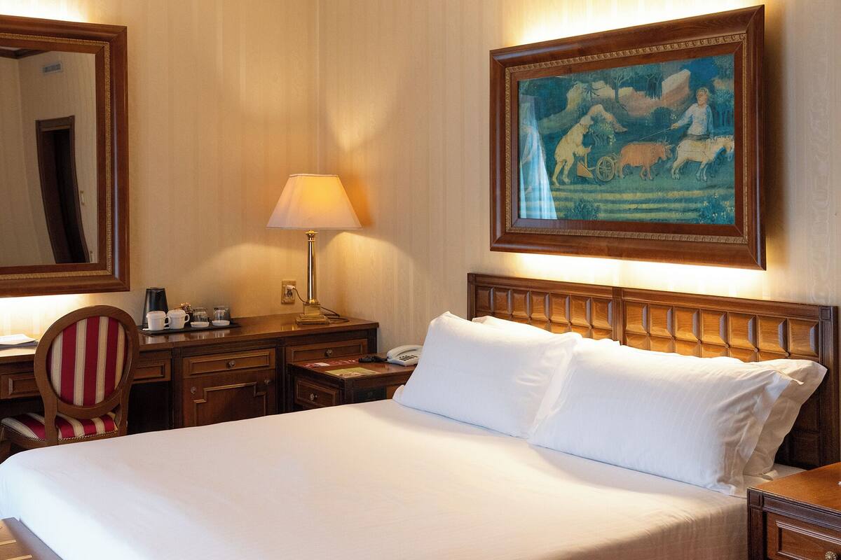 Una habitación de hotel con una cama grande, un escritorio, una silla, una mesita de noche, una lámpara, un cuadro y una alfombra.