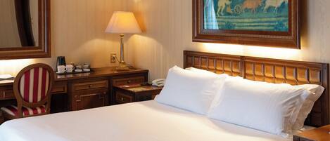 A hotel room with a large bed, a desk, a chair, a nightstand, a lamp, a painting, and a carpet.