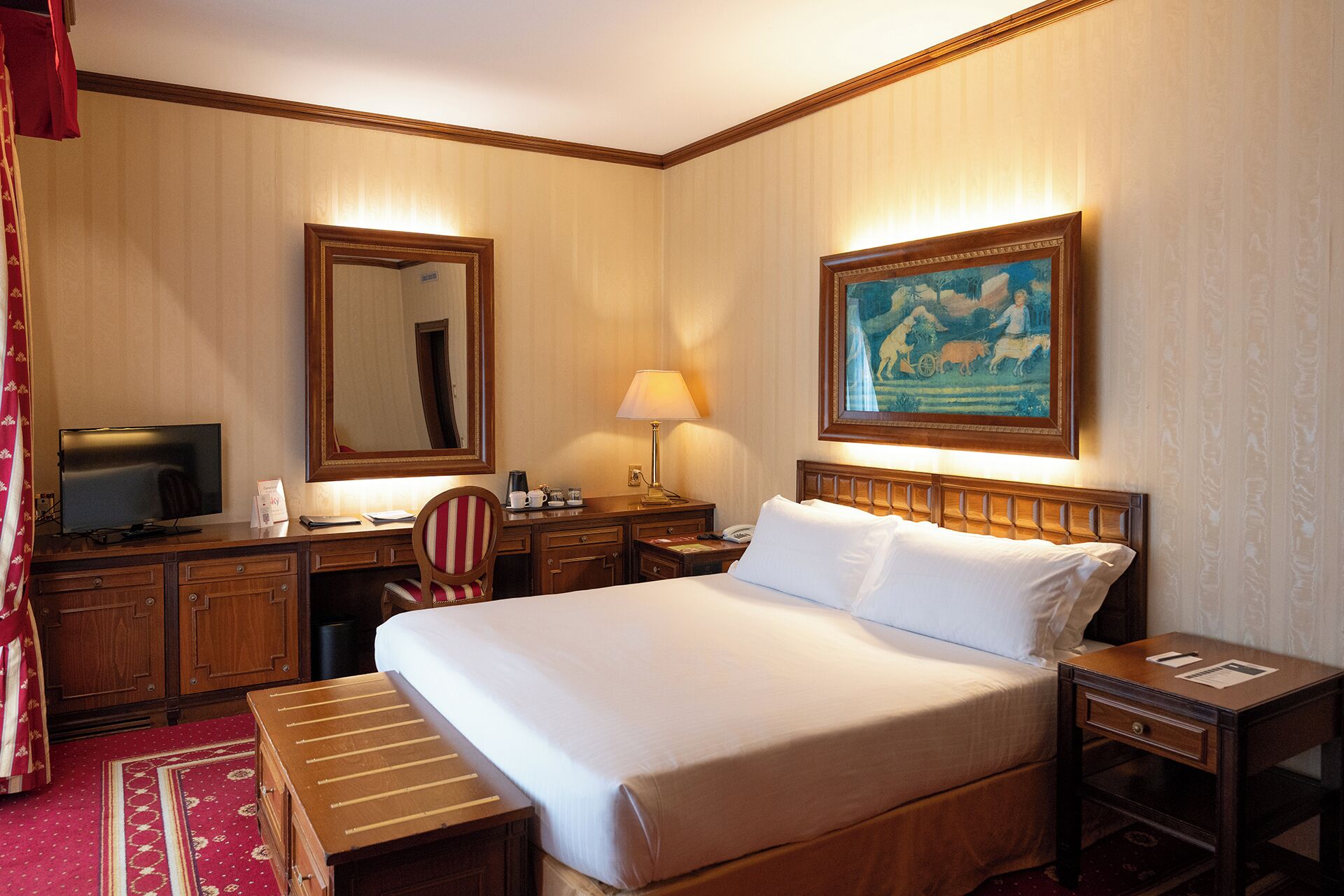 Habitación de hotel con cama, escritorio, silla, televisor, lámpara, cuadro y cortina roja.