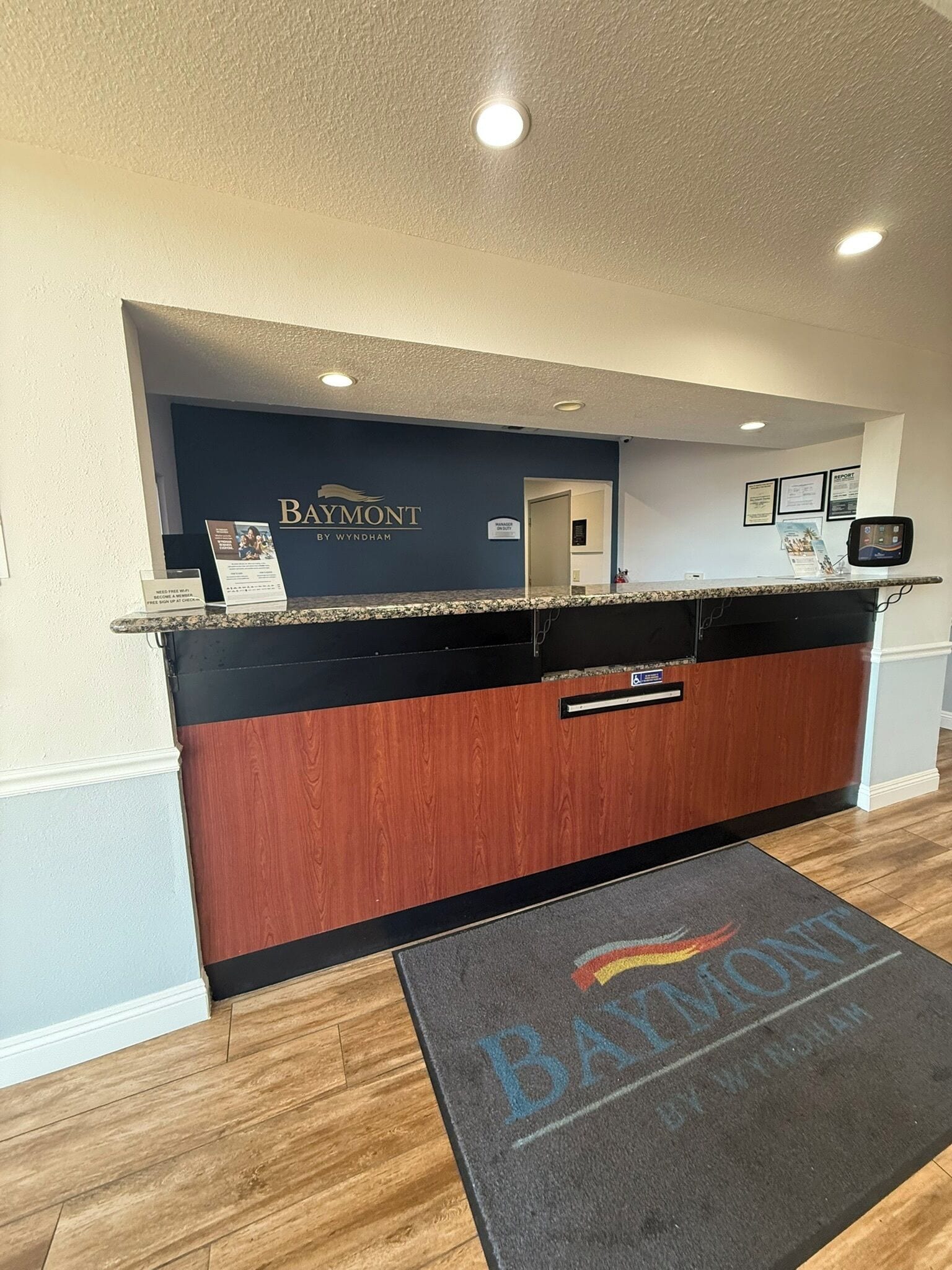 Un área de recepción con un mostrador de madera oscura, una placa con la identificación 'Baymont by Wyndham' y un felpudo con el nombre del hotel.