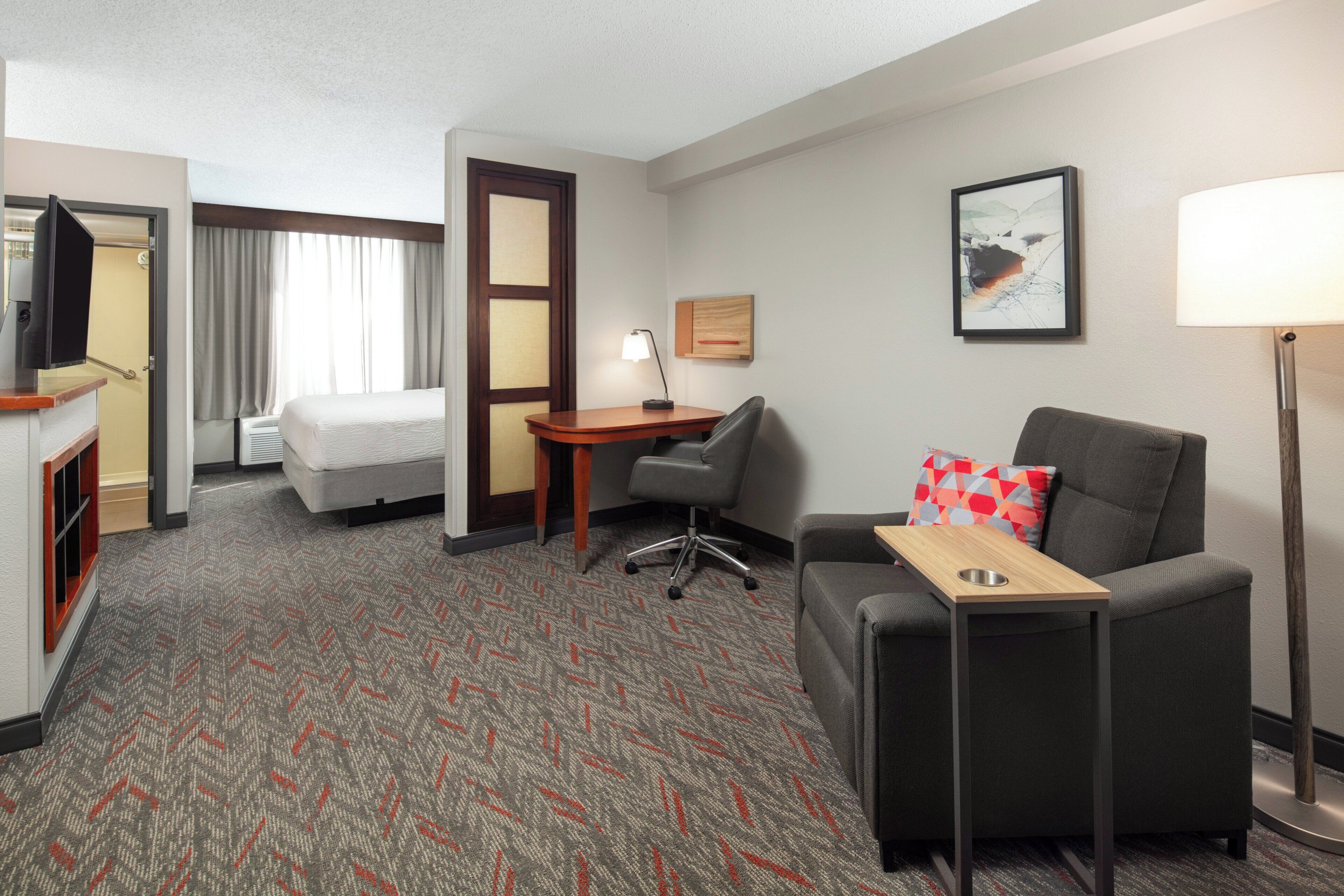 A hotel room with a bed, a desk, a chair, a sofa, a lamp, and a painting on the wall.