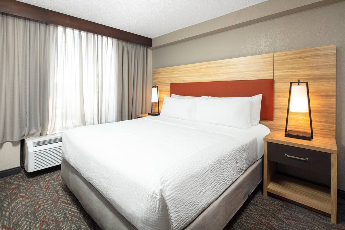 A hotel room with a large bed, a nightstand with a lamp, a heater, and a window with curtains.