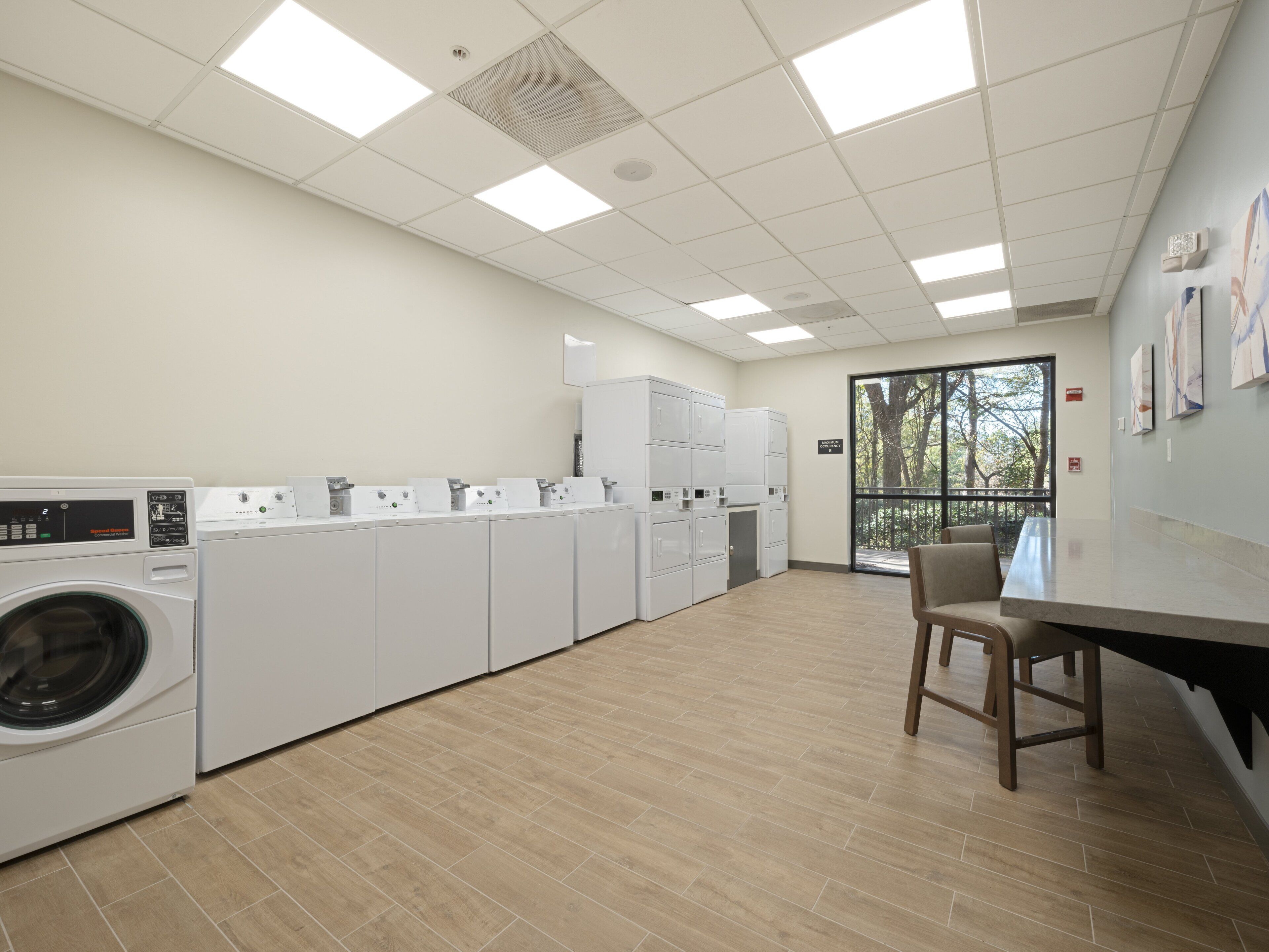 A laundry facility with multiple washing machines and dryers, a seating area, and a view of trees through a large window.