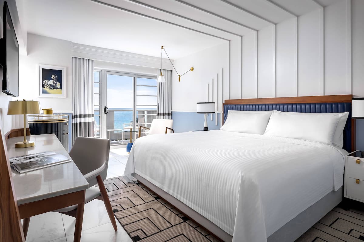 A hotel room with a large bed, a desk with a chair, a TV, and a view of the sea.