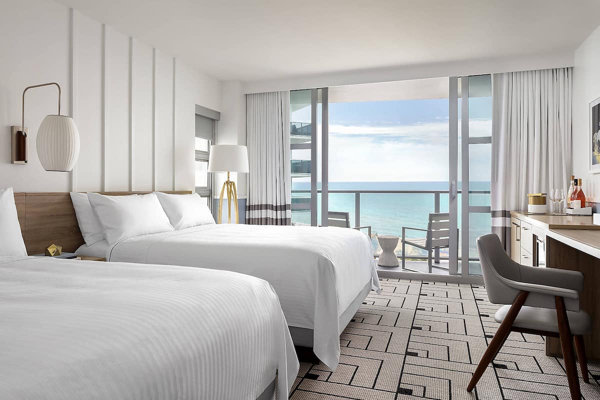 A hotel room with two beds, a desk, a chair, and a view of the ocean.