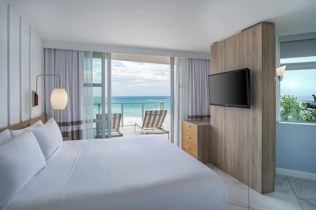 A modern hotel room with a large bed, a flat-screen TV, a wooden dresser, and a balcony with a view of the ocean.
