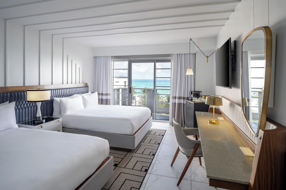 A hotel room with two beds, a desk, a TV, and a large window with a view of the ocean.