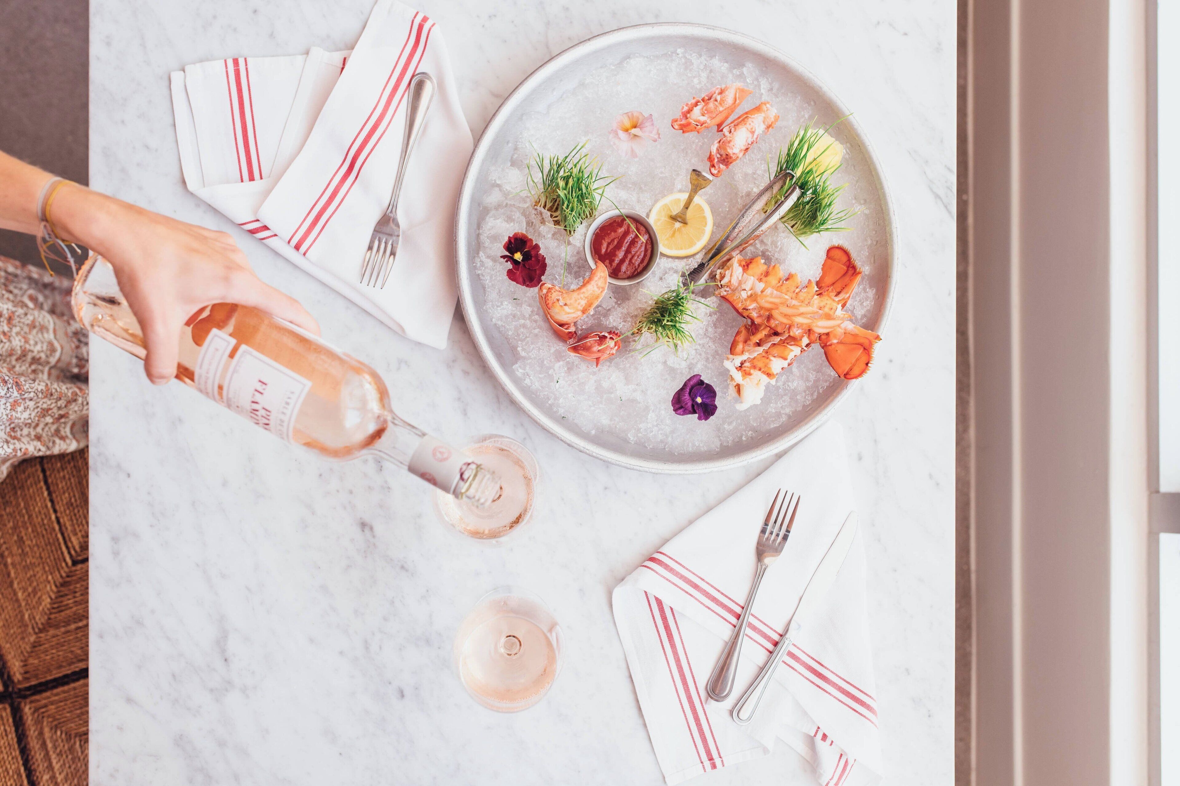 A seafood platter with shrimp and lemon on ice, garnished with herbs and flowers, and a bottle of sauce on a marble table.