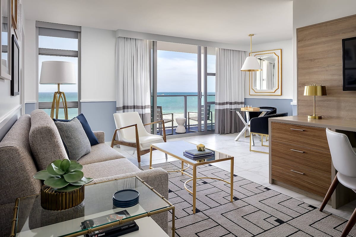 A modern hotel room with a sofa, coffee table, and dining area. There is a view of the ocean through large windows.