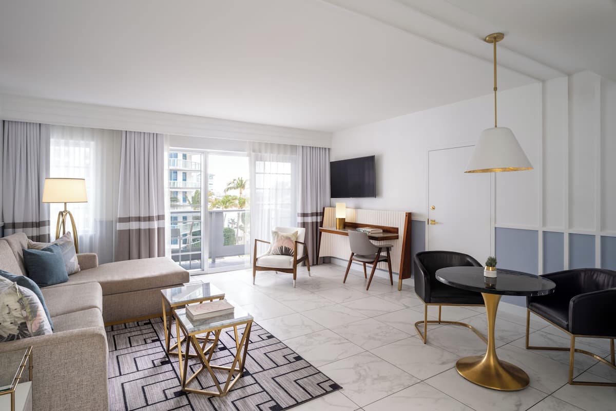 A modern hotel room with a sofa, a dining table, a desk, and a TV mounted on the wall.
