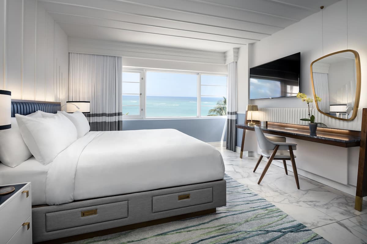 A modern hotel room with a large bed, a desk, a chair, a TV, and a view of the ocean.