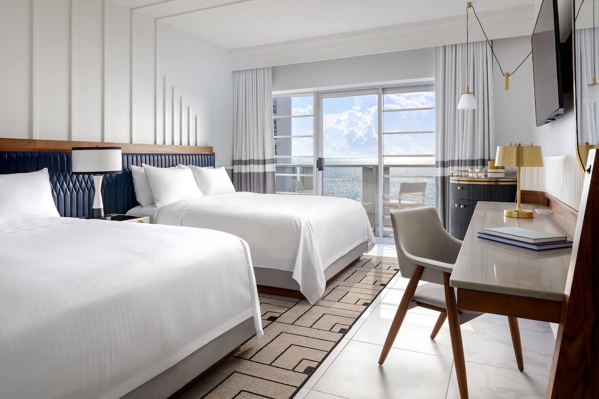 A hotel room with two beds, a desk with a chair, a TV, and a view of the sea.