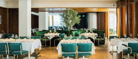 A restaurant with green booths and white tables, wooden ceiling beams, and a large tree in the center.
