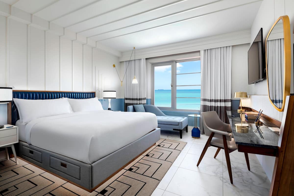 A modern hotel room with a large bed, a desk with a chair, a TV, and a view of the sea.