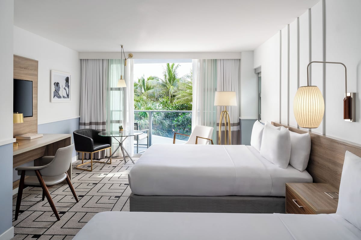 A hotel room with two beds, a desk, chairs, a TV, and a balcony with a view of palm trees.