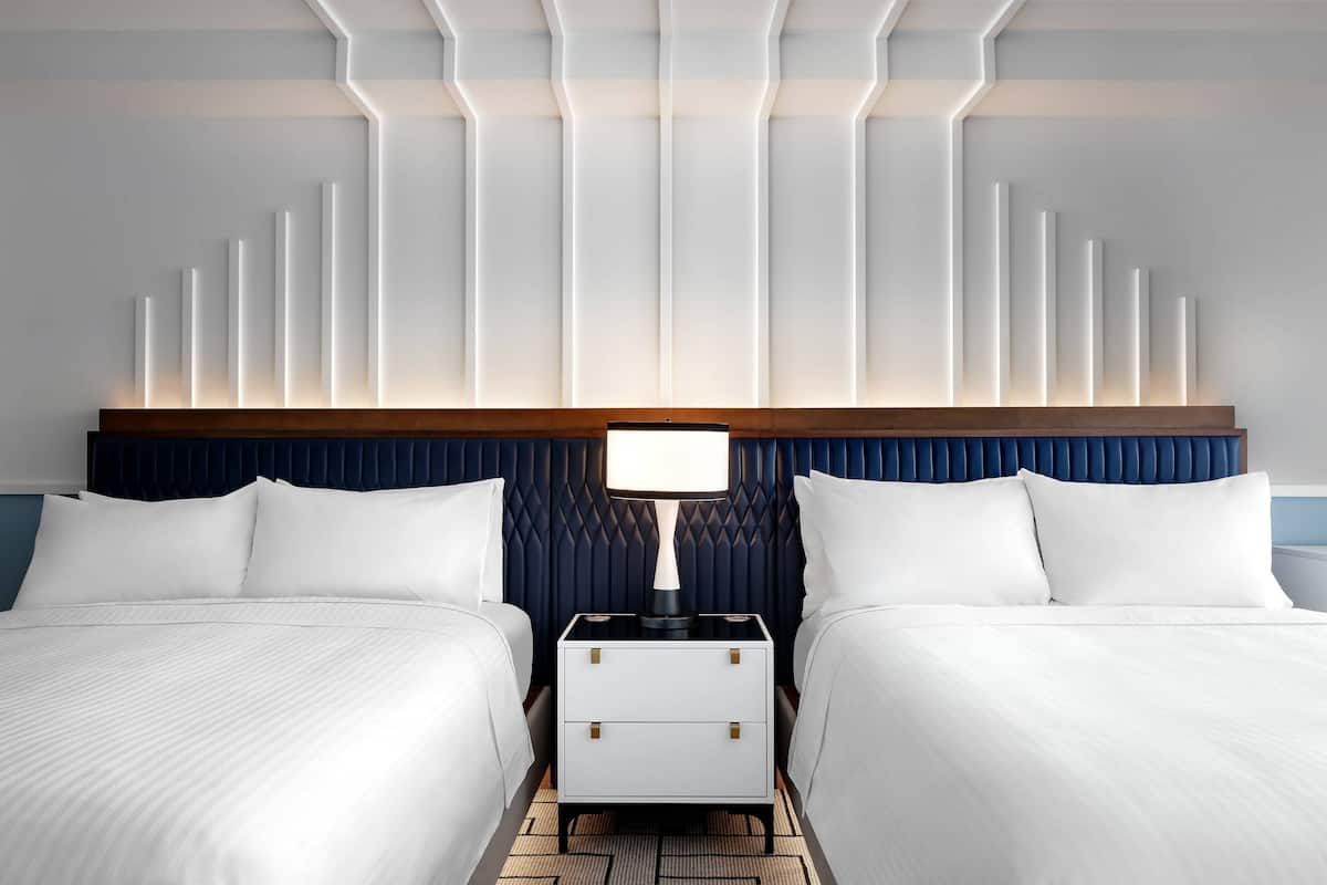 A modern hotel room with two beds, a nightstand, and a wall with vertical light panels.