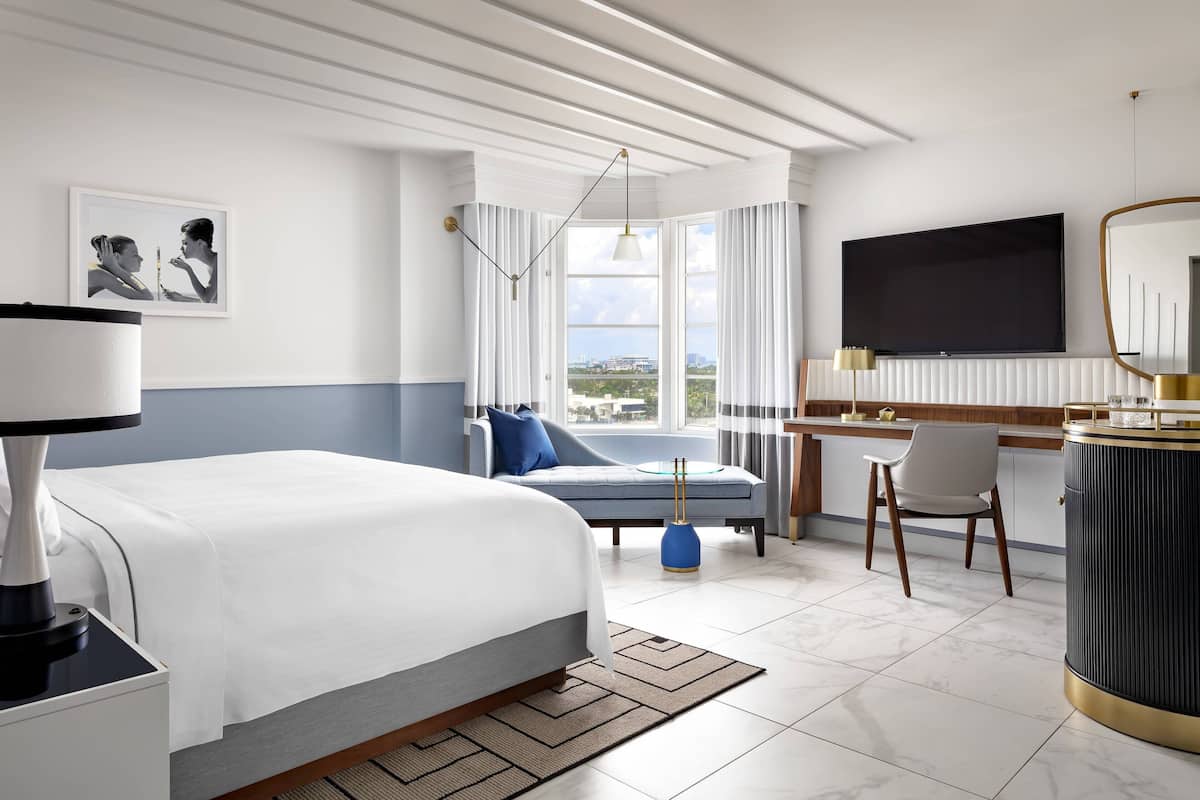 A modern hotel room with a large bed, a desk, a chair, a TV, and a view of the city.