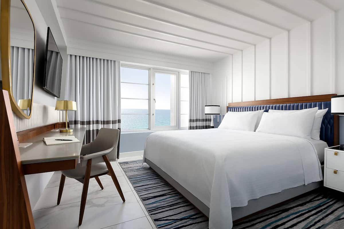 A hotel room with a large bed, a desk with a chair, a TV mounted on the wall, and a view of the sea through the windows.