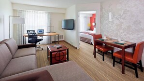 A hotel room with a sofa, a dining table with chairs, a TV mounted on the wall, and a bed in the background. - Residence Inn Lansing West (Lansing)