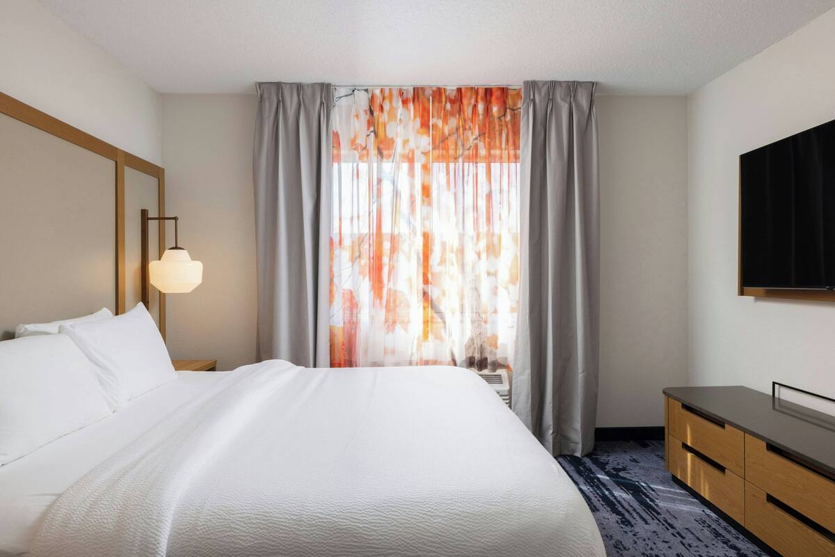 A hotel room with a large bed, a bedside table with a lamp, a dresser, and a large window with curtains.