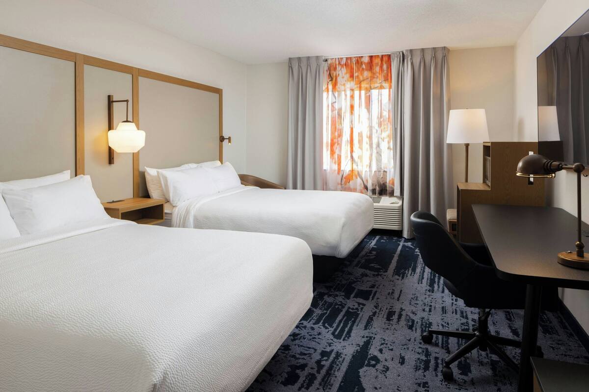 A hotel room with two beds, a desk, a chair, a lamp, and a window with curtains.