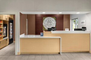 Un mostrador de recepción de hotel con un panel de pared de madera, una gran pieza de arte circular en la pared y una zona de minibar con aperitivos.