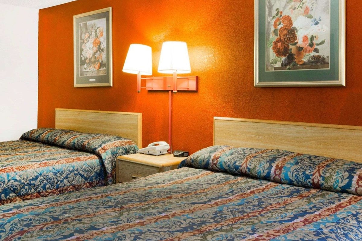 A hotel room with two beds, each with patterned bedding, a nightstand with a lamp, and framed floral artwork on the walls.