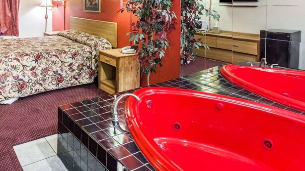 Habitación de hotel con una cama grande, una bañera roja, una mesita de noche de madera, dos lámparas fijadas a la pared y un espejo grande que refleja un televisor y un armario.