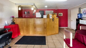 Un espace d’accueil comprenant un comptoir de réception en bois, un mur rouge, un tapis noir, une petite table recouverte d’une nappe rouge et un distributeur d’eau.