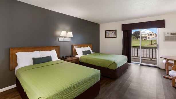 Une chambre d’hôtel avec deux lits, une tête de lit en bois, une table de chevet avec une lampe, une fenêtre donnant sur l’extérieur et une porte-fenêtre coulissante.