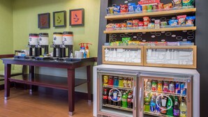 Un área de kitchenette con una cafetera, un refrigerador lleno de bebidas y estantes con bocadillos y dulces.