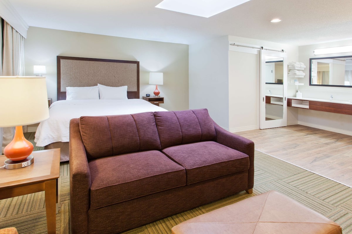 A hotel room with a bed, a sofa, a nightstand with a lamp, a bathroom with a mirror, and a shower area.