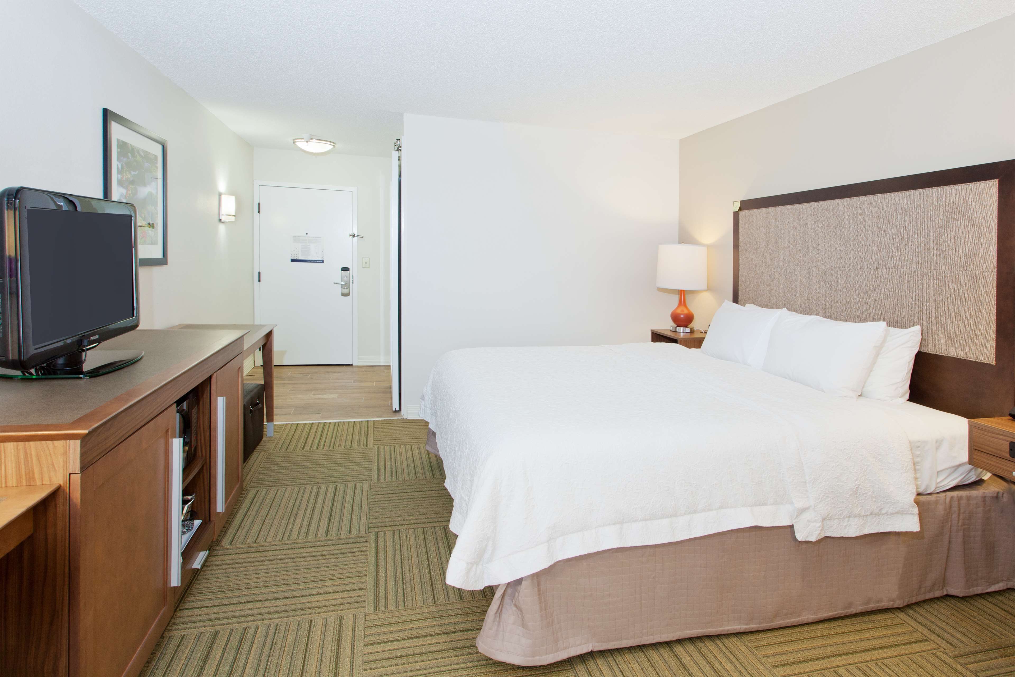 A hotel room with a bed, a desk with a television, a lamp, and a door.