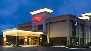 Un hotel Hampton Inn con un letrero iluminado, una entrada cubierta y un asta con la bandera estadounidense.