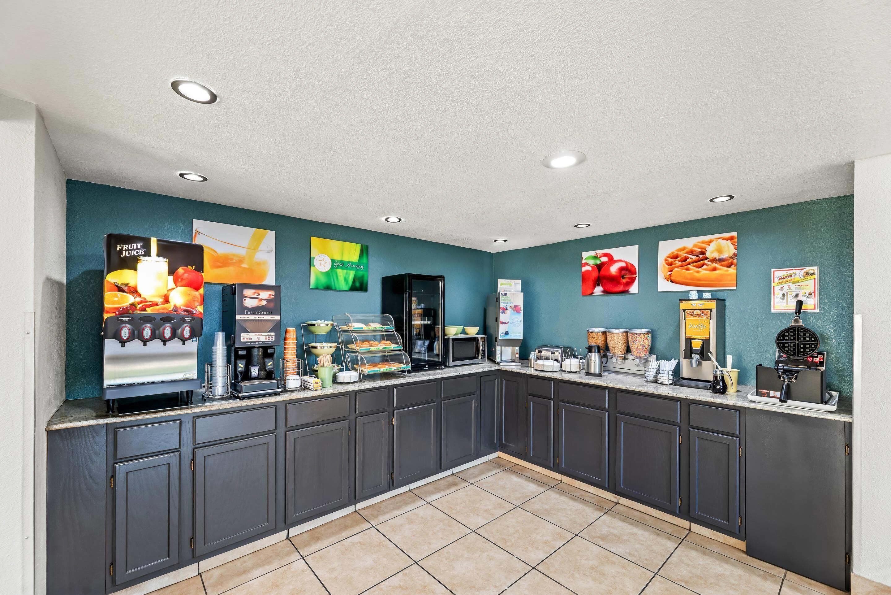 A self-service breakfast area with a juice dispenser, coffee machine, and food display.