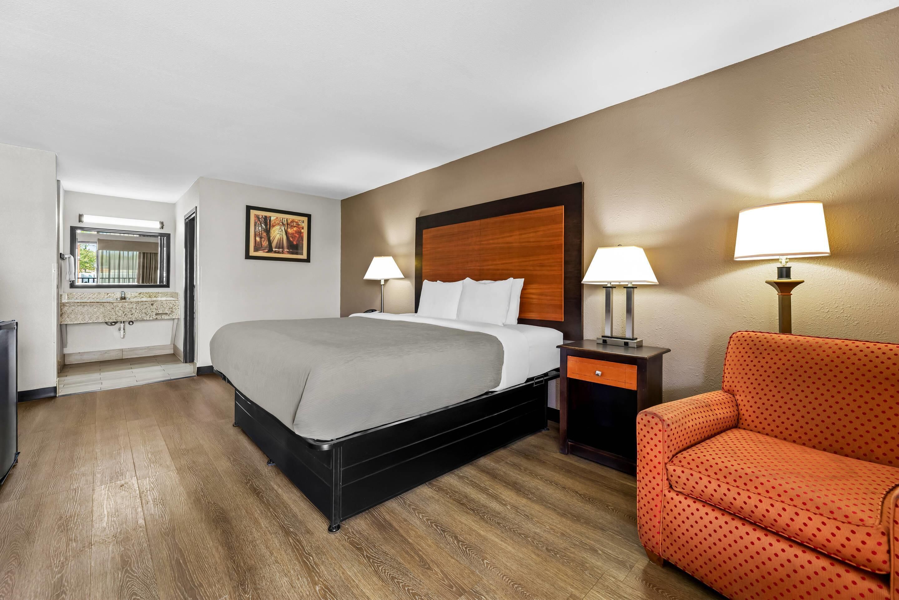 A hotel room with a bed, two bedside lamps, a sofa, a small table, and a bathroom visible through an open door.