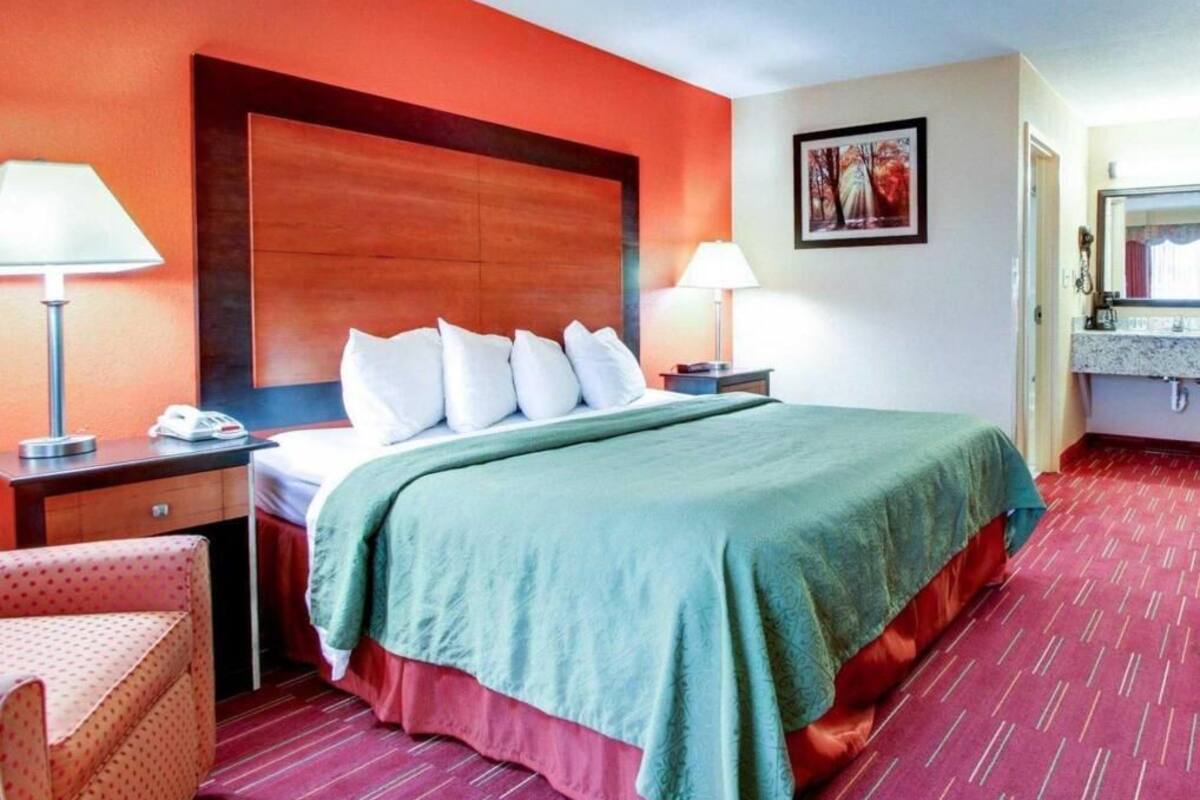 A hotel room with a bed, two bedside tables, a chair, a desk, a lamp, a painting, and a bathroom with a sink and mirror.