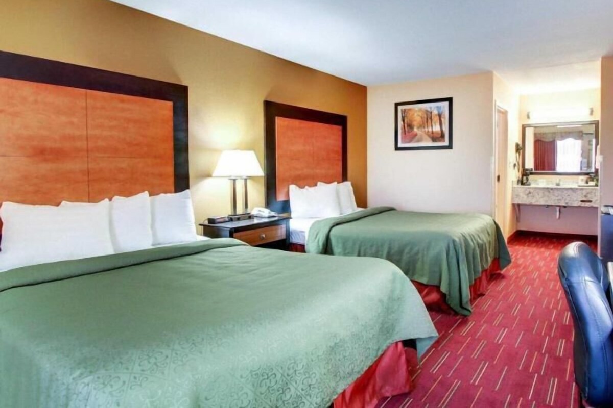 A hotel room with two beds, a nightstand with a lamp, a painting on the wall, and a bathroom with a mirror and sink.