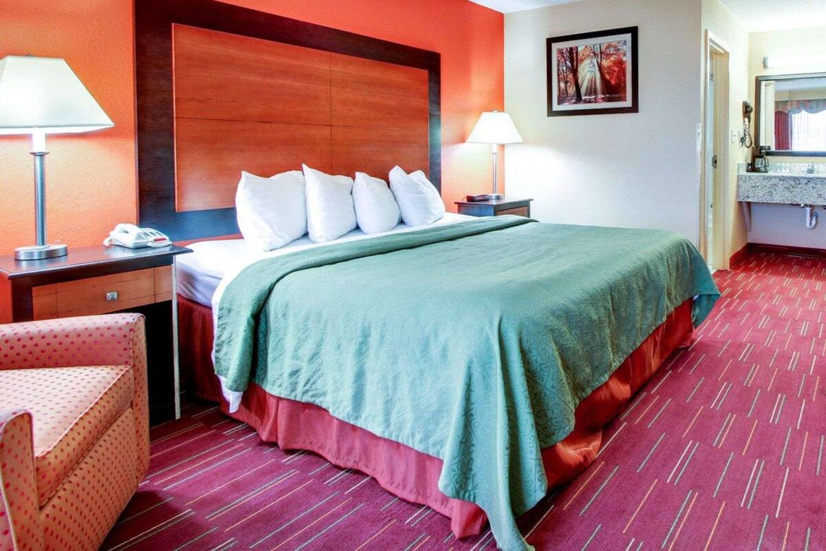A hotel room with a large bed, two bedside tables with lamps, a chair, a bathroom with a sink and mirror, and a framed picture on the wall.
