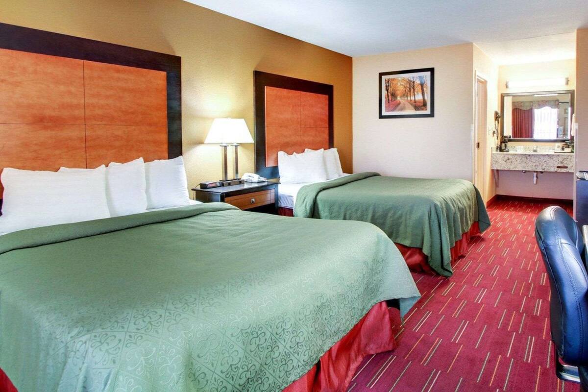 A hotel room with two beds, a nightstand with a lamp, a painting on the wall, and a bathroom with a mirror and sink.