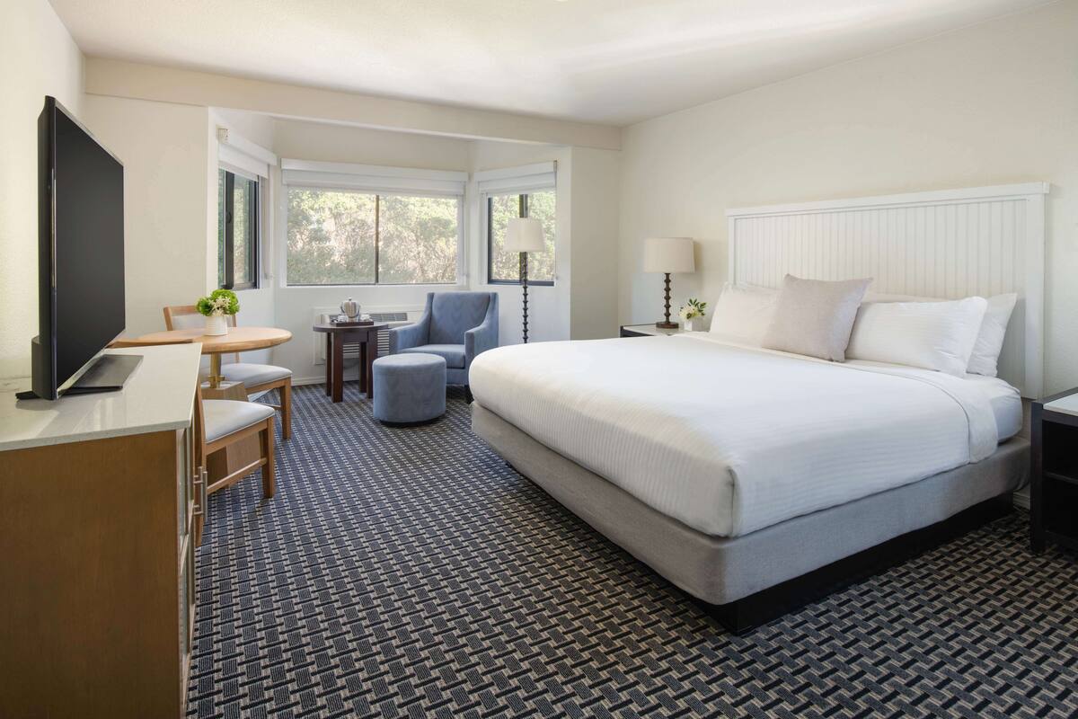 A hotel room with a bed, a desk with a TV, a chair, a small table, and a lamp.