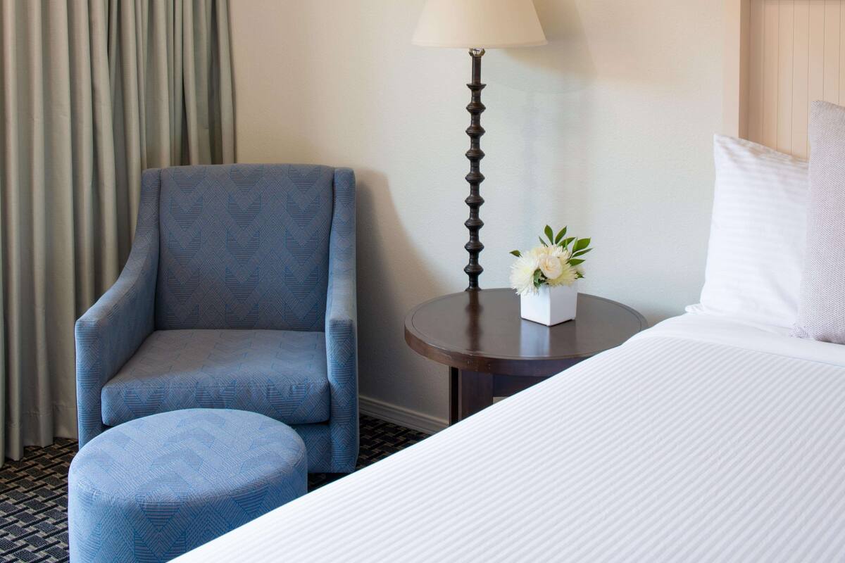 A hotel room with a bed, a chair, a small table with a lamp, and a vase with flowers.