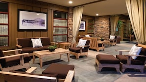 A cozy lounge area with wooden furniture, stone walls, and large windows with curtains.