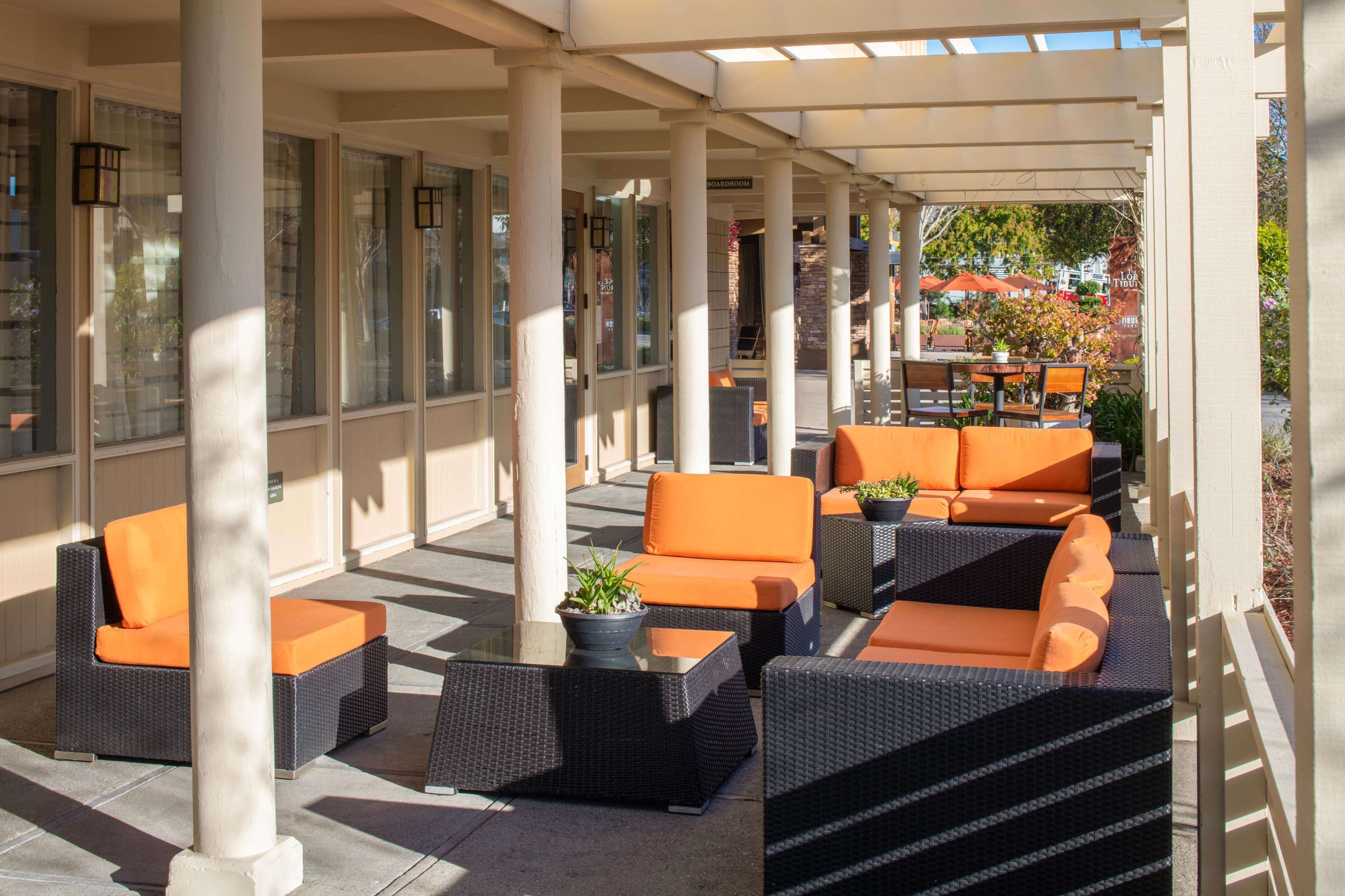 An outdoor seating area with wicker furniture and orange cushions, featuring a glass wall and columns.
